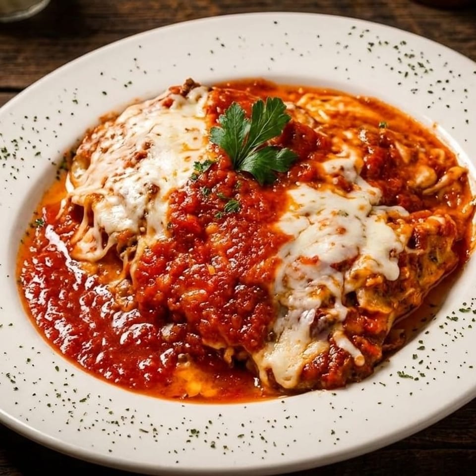 A white plate holds a serving of lasagna topped with melted cheese, red sauce, and a parsley garnish.