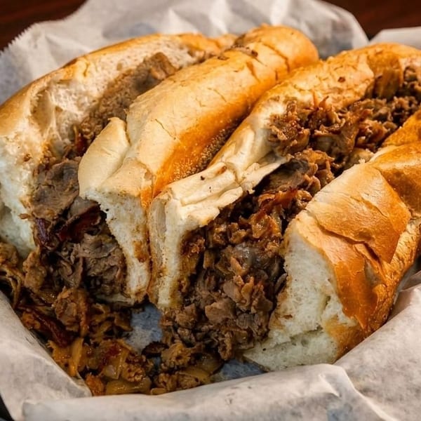 Two hearty Philly cheesesteak sandwiches, overflowing with meat and onions, wrapped in white paper.